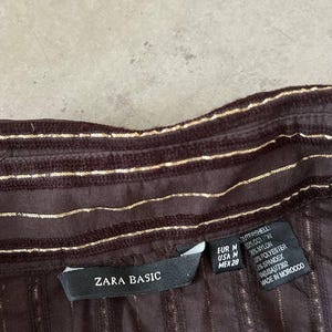 May include: Close-up of a brown shirt with vertical gold stripes. The shirt has a collar and a label that reads "ZARA BASIC". The fabric is a blend of cotton, nylon, polyester, and spandex.