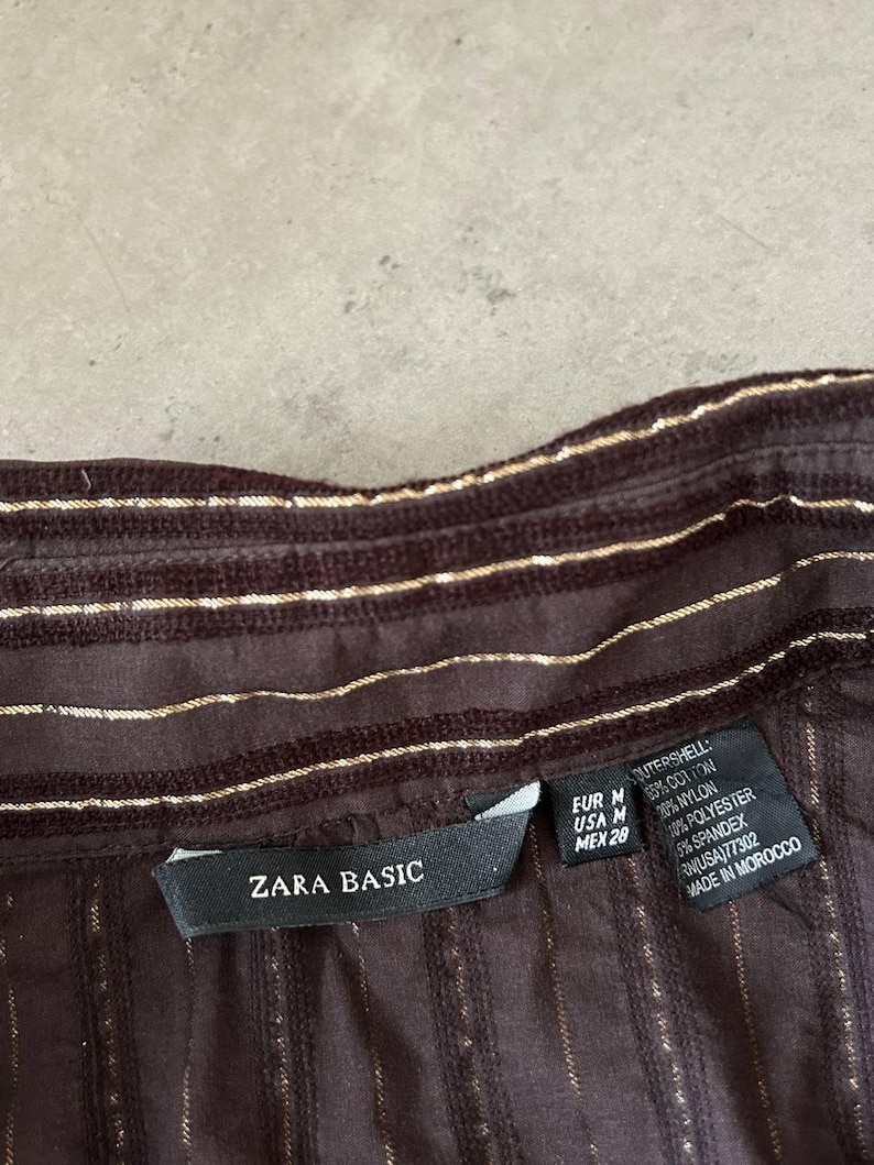 May include: Close-up of a brown shirt with vertical gold stripes. The shirt has a collar and a label that reads "ZARA BASIC". The fabric is a blend of cotton, nylon, polyester, and spandex.