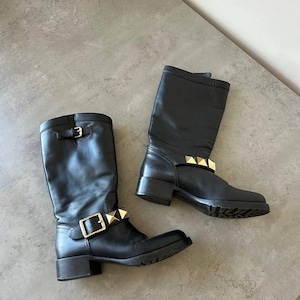Peut inclure: Une paire de bottes noires en cuir jusqu'aux genoux avec des d&eacute;tails m&eacute;talliques dor&eacute;s. Les bottes sont dot&eacute;es d'une fermeture &agrave; boucle et de clous d&eacute;coratifs. Les semelles sont noires.