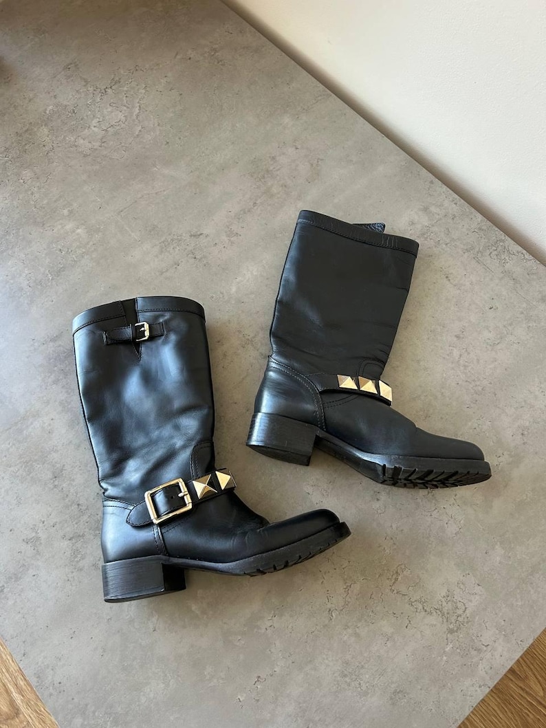 Peut inclure: Une paire de bottes noires en cuir jusqu'aux genoux avec des d&eacute;tails m&eacute;talliques dor&eacute;s. Les bottes sont dot&eacute;es d'une fermeture &agrave; boucle et de clous d&eacute;coratifs. Les semelles sont noires.