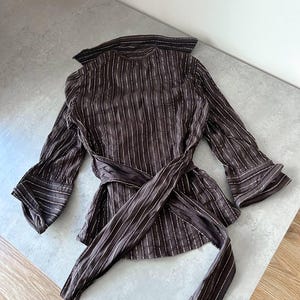 May include: A brown, long-sleeved blouse with vertical white stripes. The blouse features a wrap-around tie at the waist and flared sleeves. The collar and cuffs have a contrasting trim. The fabric appears lightweight and flowing.