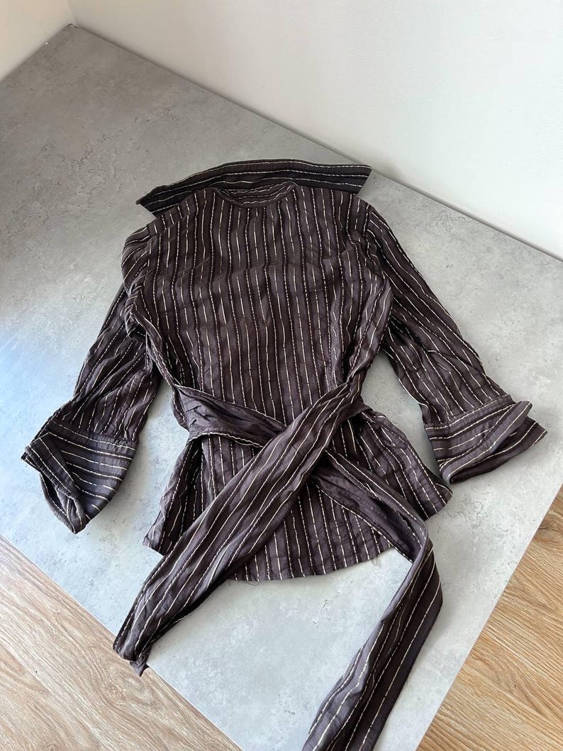 May include: A brown, long-sleeved blouse with vertical white stripes. The blouse features a wrap-around tie at the waist and flared sleeves. The collar and cuffs have a contrasting trim. The fabric appears lightweight and flowing.