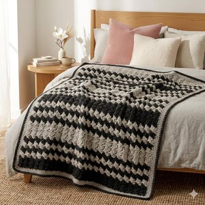 May include: A crochet blanket with a geometric pattern in black and grey, draped over a bed. The blanket's design features a repeating square pattern. The bed is in a room with neutral tones, a wooden side table, and decorative pillows.