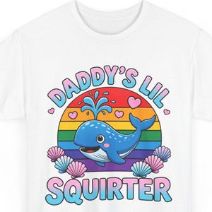 Könnte beinhalten: Ein weißes T-Shirt mit kurzen Ärmeln und einer Grafik eines blauen Wals, der Wasser spritzt, umgeben von rosa und blauen Muscheln und kleinen Herzen. Hinter dem Wal ist eine regenbogenfarbene Sonne. Der Text DADDY'S LIL SQUIRTER ist blau.