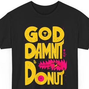 Può includere: Una t-shirt nera a maniche corte con una grande scritta gialla che recita GOD DAMNIT DONUT. Il design include una piccola icona a forma di trofeo, un ananas, tre icone di teschi e un elemento grafico rosa che raffigura una persona che fugge da una creatura.