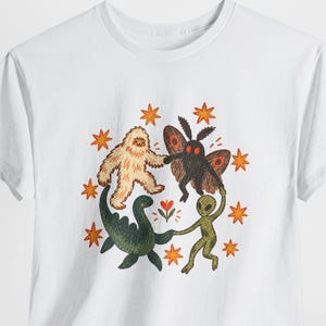 Può includere: Una t-shirt bianca a maniche corte con una grafica centrale raffigurante uno yeti beige, una falena scura, una creatura marina verde e un alieno verde che si tengono per mano in cerchio attorno a un piccolo fiore, circondati da stelle arancioni.