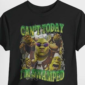 Peut inclure: Un t-shirt noir à manches courtes avec une impression graphique du personnage Shrek dans diverses poses, dont une avec des lunettes de soleil violettes. Le texte CAN'T TODAY I'M SWAMPED est imprimé en vert usé au-dessus et en dessous.