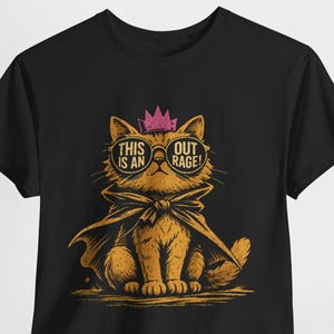 Op de afbeelding: Een zwart t-shirt met een afbeelding van een oranje kat die een roze kroon, een cape en een ronde zonnebril draagt. De glazen van de bril bevatten de tekst THIS IS AN OUTRAGE! De kat is afgebeeld in een zittende houding met een serieuze blik.