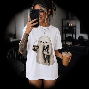 Op de afbeelding: Een wit T-shirt met korte mouwen voorzien van een grafische illustratie van een geest die een dampende bruine mok met de tekst MEH vasthoudt, terwijl hij ook een kleine zwart-witte Boston Terrier in zijn armen houdt.