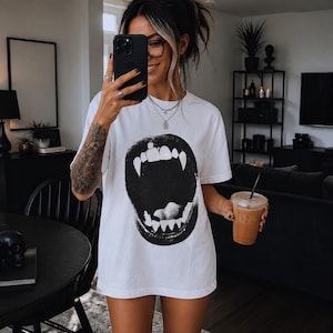 Pode incluir: Uma t-shirt branca de manga curta com uma grande estampa gráfica preta centralizada de uma boca aberta com presas proeminentes. O design é ousado e de alto contraste, cobrindo a área do peito da peça. A t-shirt tem gola redonda e um corte relaxado e solto. O tecido parece ser de algodão padrão e o gráfico está impresso com uma tinta preta sólida e opaca.