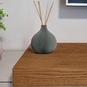 Può includere: Un vaso diffusore grigio con un motivo a spirale e diversi bastoncini di legno. Il vaso è appoggiato su una superficie di legno. È un oggetto decorativo per la profumazione della casa.