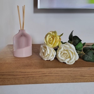 Può includere: Un diffusore rosa con bastoncini di legno e tre rose artificiali, una gialla e due bianche, su una superficie di legno.