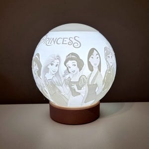 Può includere: Una lampada sferica bianca con immagini incise di principesse Disney. La parola "PRINCESS" è sopra le immagini delle principesse. La lampada poggia su una base circolare marrone. La lampada è illuminata dall'interno, creando una luce soffusa.