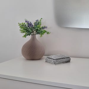 Könnte beinhalten: Eine hellbraune, spiralförmige Keramikvase mit einem Strauß lila und grüner Blumen steht auf einer weißen Oberfläche neben einer silbernen, mit Schmuck besetzten Schachtel.