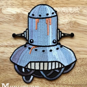May include: A blue and gray embroidered iron-on patch of a robot with orange dripping paint. The robot has a round head with a dome shape and a large, round body. The robot has two small arms and a single antenna.