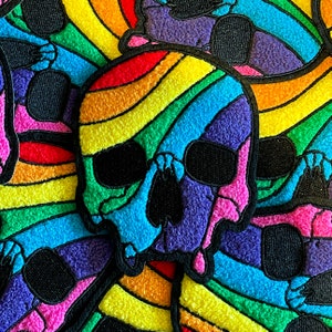 Può includere: Una toppa a forma di teschio arcobaleno con una texture pelosa. Il teschio è contornato di nero e ha un motivo arcobaleno.