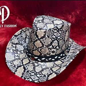 Puede incluir: Sombrero de vaquero con estampado de piel de serpiente en negro, beige y blanco. El sombrero tiene una banda negra con costuras decorativas y tachuelas plateadas. El texto "DONGY FASHION" es visible en el lado izquierdo.