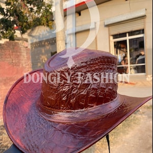Könnte beinhalten: Ein brauner Cowboyhut aus Leder mit einem strukturierten, krokodilartigen Muster. Der Hut hat eine breite Krempe und eine strukturierte Krone. Der Text "DONGLY FASHION" ist auf dem Hut aufgedruckt. Der Hut hat einen Kinnriemen.