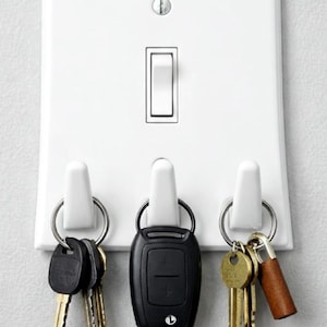 Könnte beinhalten: Weißer Schlüsselhalter im Design eines Lichtschalters, mit drei Haken für Schlüssel. Die Schlüsselanhänger halten verschiedene Schlüssel, darunter Autoschlüssel und Hausschlüssel. Der Schlüsselhalter ist an einer hellgrauen Wand montiert.