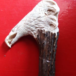 Puede incluir: Una escultura tallada de dos cabezas de águila, hecha de un material de color claro, posiblemente hueso o marfil. La talla detallada muestra los picos, ojos y plumas de las águilas. La escultura está montada sobre una base de madera oscura y texturizada, sobre un fondo rojo.