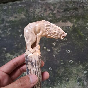 Puede incluir: Escultura tallada de un oso en posición de pie. El oso es de color marrón claro con detalles de pelaje y rasgos faciales. La escultura está montada sobre una base texturizada de color marrón oscuro, que se asemeja a un tronco de árbol.