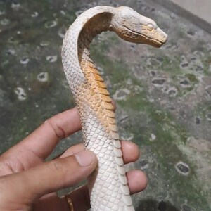 May include: A carved cobra snake figurine. The sculpture is primarily off-white with tan accents on the neck and head. The snake is posed in a striking, upright position, with detailed scale patterns.