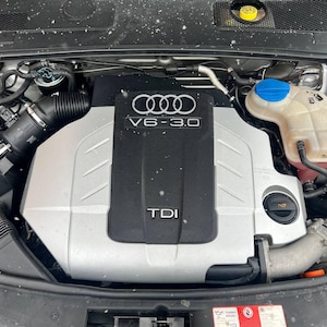 Könnte beinhalten: Ein Audi V6 3.0 TDI Motorraum mit einer zentralen schwarzen und silbernen Motorabdeckung, die das Audi-Logo, V6-3.0 und den TDI-Schriftzug trägt. Der Motorraum enthält einen durchsichtigen Kühlmittelbehälter mit blauem Deckel und einen Öldeckel.