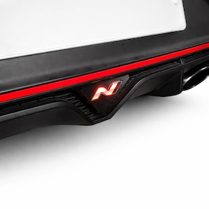 Può includere: Primo piano di un paraurti nero per auto con una striscia rossa e un logo "N" rosso incandescente. Il paraurti presenta anche una targa bianca e un diffusore inferiore nero.