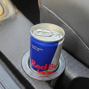 Könnte beinhalten: Eine blaue und silberne Red Bull Energy-Drink-Dose in einem Getränkehalter im Auto. Die Dose hat das Red Bull-Logo in Rot und Weiß. Der Getränkehalter ist silberfarben und der Innenraum des Autos ist schwarz.