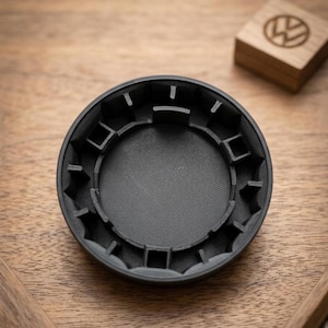 Op de afbeelding: Een zwart, cirkelvormig object met een textuur aan de binnenkant en een reeks vierkante en rechthoekige uitsparingen langs de rand. Het object staat op een houten oppervlak, met een klein houten blok met een logo op de achtergrond.