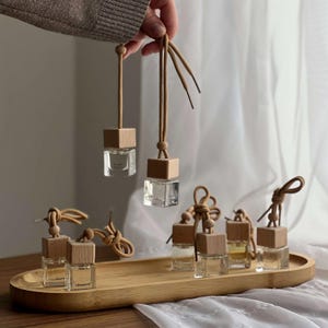Op de afbeelding: Transparante glazen auto-geurverfrissers met houten tops en bruine koorden worden gepresenteerd op een houten dienblad. Twee worden door een hand omhoog gehouden. De luchtverfrissers hebben een vierkante vorm en zijn gevuld met een heldere vloeistof.