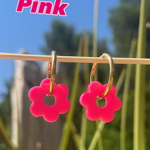 May include: A pair of gold-toned hoop earrings featuring bright pink flower-shaped charms. The word Pink is printed in a bold, white font with a pink outline in the upper left corner of the image. The earrings are suspended from a thin horizontal rod.