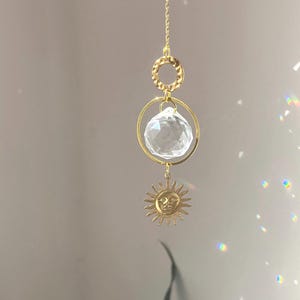 Peut inclure: Un attrape-soleil décoratif avec une boule de cristal transparente et un charm en forme de soleil doré. Le soleil a un visage souriant et des rayons. Le capteur solaire est suspendu par une chaîne dorée et possède un anneau doré.