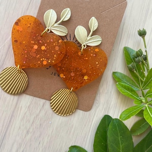Puede incluir: Un par de pendientes en forma de corazón de resina naranja translúcida con purpurina dorada incrustada. Cada corazón está rematado con un dije de hoja de metal dorado y un disco circular de metal dorado con textura estriada.