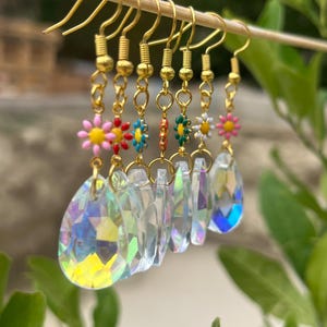 Könnte beinhalten: Eine Sammlung von sechs Hängeohrringen, die an einem Holzstab hängen. Jeder Ohrring besteht aus einem goldfarbenen Haken, einem kleinen bunten Emaille-Blumenanhänger in Pink, Rot, Blau, Orange, Grün oder Weiß und einem klaren, irisierenden Tropfenkristall.