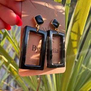 May include: A pair of dangle earrings featuring black rectangular frames suspended from smaller black diamond-shaped studs with gold-tone metal accents. The earrings are attached to a brown cardboard display card with the text By iiia printed.
