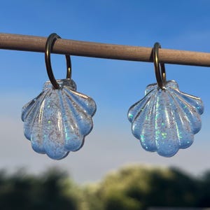 Könnte beinhalten: Ein Paar Creolen mit durchscheinenden, muschelförmigen Anhängern. Die Anhänger sind klar und mit eingebettetem, irisierendem Glitzer versehen, der das Licht reflektiert. Sie hängen an einfachen, dünnen Creolen aus dunkel getöntem Metall.