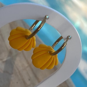 Op de afbeelding: Een paar gouden oorringen met heldergele schelpenbedels. De oorbellen worden gepresenteerd op een witte standaard tegen een vage blauw-witte achtergrond. De schelpenbedels hebben een gestructureerd, geribbeld ontwerp.