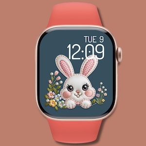 Op de afbeelding: Een smartwatch met een koraalkleurige band en een goudkleurige behuizing. De wijzerplaat toont een geborduurd konijn met roze ruitjesoren en bloemmotieven. De tijd is 12:09 uur op dinsdag.