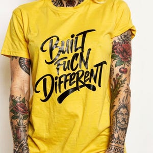Könnte beinhalten: Gelbes T-Shirt mit dem schwarzen Schriftzug "Built Fucn Different". Das kurzärmelige T-Shirt wird von einer Person mit Tätowierungen an Armen und Hals getragen.
