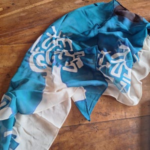 May include: A flowing, lightweight scarf in shades of turquoise, white, and cream. The scarf features a repeating abstract design in white against a turquoise background. The scarf is draped on a wooden surface, showcasing its delicate texture and vibrant colors.