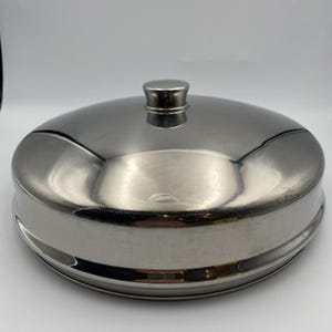 May include: A stainless steel, round food cover with a small handle on top. The cover has a shiny, reflective surface and a slightly domed shape. The base of the cover has a raised edge. The overall design is simple and functional.