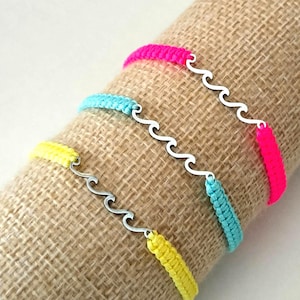 May include: Three colourful bracelets with a wave design. The bracelets are made of woven neon pink, blue, and yellow threads, connected by a silver-coloured wave-shaped charm. The bracelets are displayed on a tan fabric.