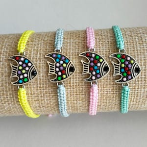 Puede incluir: Cuatro pulseras coloridas con forma de pez. Cada pulsera presenta un colgante de pez plateado con detalles de puntos de colores y un ojo negro. Las pulseras tienen cordones tejidos amarillos, celeste, rosa y turquesa.