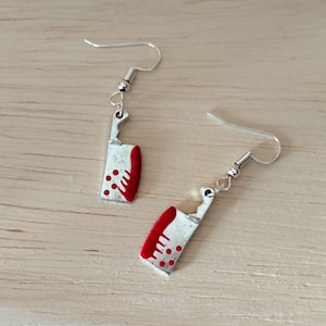 Puede incluir: Un par de pendientes colgantes plateados con forma de cuchilla en miniatura. Las hojas están pintadas de rojo con pequeños puntos rojos, y los mangos son plateados. Los pendientes cuelgan de ganchos plateados.