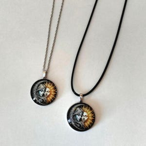May include: Two round pendant necklaces with a celestial design. One pendant hangs from a silver chain, the other from a black cord. The pendants show a woman's face with the sun and moon, in yellow, blue, and black.