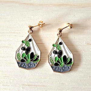 Könnte beinhalten: Ein Paar goldfarbene Ohrstecker. Jeder Ohrring hat ein weißes, geometrisches Terrariendesign mit grünen und schwarzen Kaktusdetails. Die Ohrringe haben einen Stecker-und-Kugel-Verschluss, perfekt für einen Hauch von Natur.