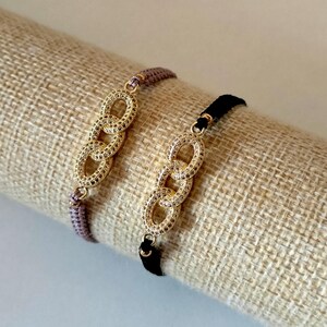 Könnte beinhalten: Zwei Armbänder mit goldfarbenen Kettengliedern, die mit kleinen klaren Steinen besetzt sind. Ein Armband hat ein hellviolettes Band, das andere ein schwarzes Band. Die Glieder sind in einer vertikalen Reihe in der Mitte angeordnet.