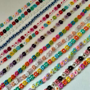 Puede incluir: Filas de cadenas de margaritas coloridas en varios colores, incluyendo rosa, naranja, amarillo, verde y morado. Cada margarita tiene un centro blanco o dorado, y las cadenas están dispuestas diagonalmente.