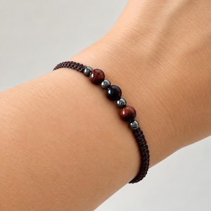 May include: A brown braided cord bracelet featuring a central arrangement of two reddish-brown tiger eye beads, one solid black bead, and four small metallic grey hematite beads spaced evenly along the woven band for a simple, adjustable design.